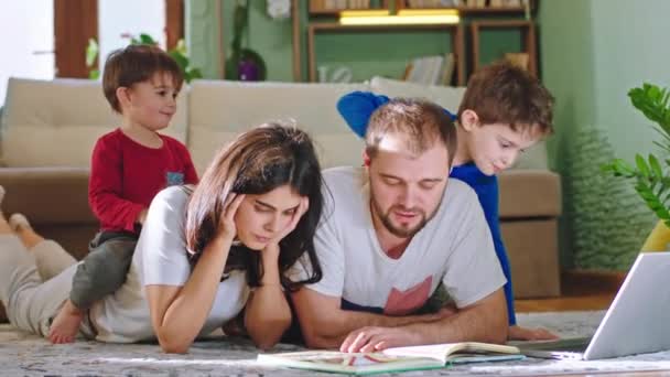 Glückliche Familie, die Zeit zu Hause verbringen, lesen sie Geschichten Buch lächelnd groß ihre beiden Kinder spielen um sie herum legen sie sich auf den Boden — Stockvideo