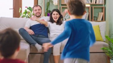 Gülümseyen anne ve baba kanepede oturup koşan çocuklarını bekliyorlar. Birbirlerine sarılıyorlar ve birlikte komik oyunlar oynuyorlar.