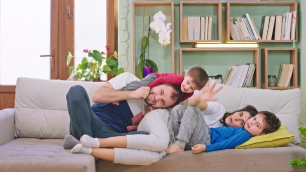 Atractiva familia joven mirando directamente a la cámara disfrutando del tiempo con sus hijos que yacen en el sofá y jugando divertido juntos — Vídeos de Stock