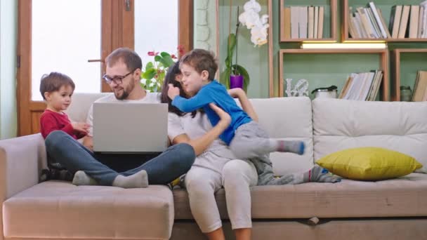 Charismatische Familie verbringt Zeit zu Hause Papa arbeitet von zu Hause aus an seinem Laptop, während Mama auf dem Sofa sitzt und mit ihrem großen Sohn spielt — Stockvideo