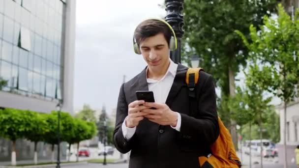 Красивий чоловік з чорним костюмом і зеленими навушниками бере свій телефон, щоб набрати щось зосереджене і з щасливою посмішкою — стокове відео