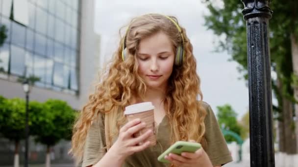 Mujer guapa con un pelo largo rizado escuchando música de los auriculares beber un poco de café y charlar con alguien en el teléfono inteligente. 4k — Vídeo de stock