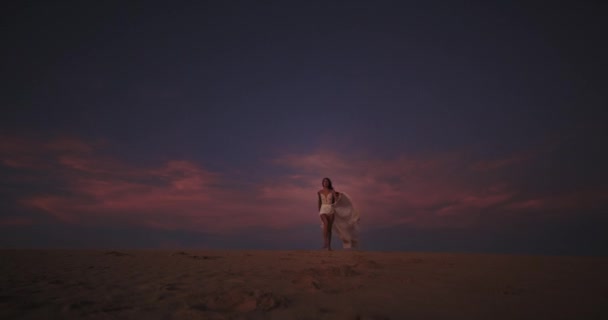 Romantische dame met een lange witte jurk die door het witte zand loopt in een prachtig zonsondergang landschap. 4k — Stockvideo