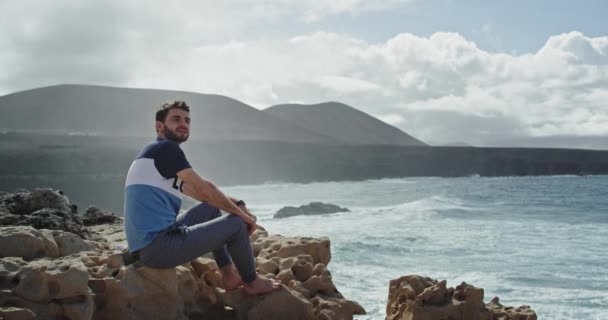 Fiatalember a csodálatos táj közepén, kilátással a tengerre és a hegyre, egy fickó ül a sziklán, és élvezi a kilátást. — Stock videók