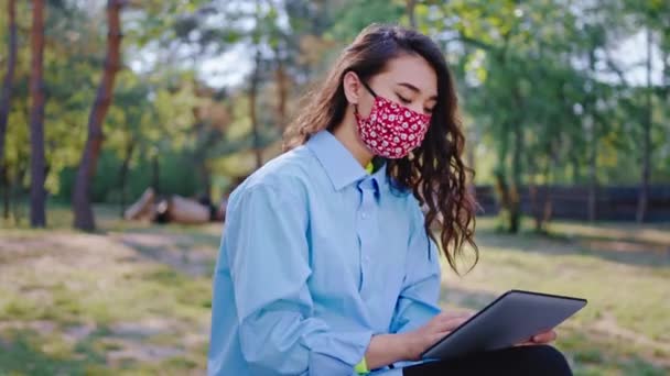 Signora asiatica con i capelli lunghi e maschera protettiva in quarantena seduta in mezzo al parco prendere la tavoletta elettronica per studiare al concetto di aria fresca di Coronavirus. Girato su ARRI Alexa Mini — Video Stock