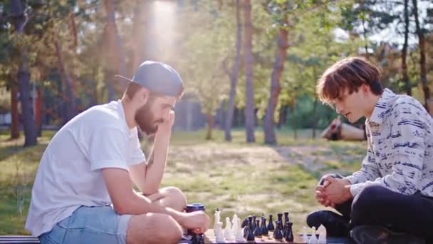 Zwei befreundete charismatische Typen spielten konzentriert auf einem Schachspiel inmitten eines großen grünen Parks, während sie auf dem Stuhl saßen. 4k — Stockvideo