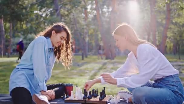 Charismatische dames met een mooie glimlach in het midden van een groot park spelen op een schaakspel — Stockvideo