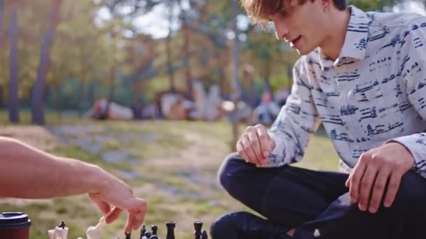 Добре виглядають молоді хлопці, які грають зосереджені на шаховій грі посеред великого зеленого парку — стокове відео