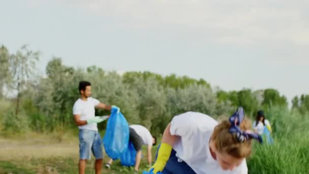 Kelompok relawan multirasial dengan senyum lebar senang membersihkan gosok dan mengumpulkan kantong plastik biru di pantai hijau besar di samping danau mereka mengenakan sarung tangan pelindung kuning — Stok Video