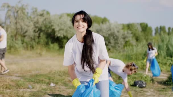 En la playa junto al lago grupo de diversos amigos voluntarios recogiendo la basura de la playa y recogiendo en las bolsas de plástico azul señora parada delante de la cámara y mirando directamente — Vídeo de stock