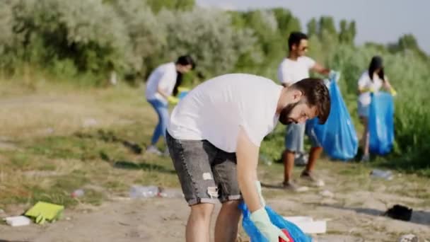 Nastoletnia grupa wolontariuszy pracująca razem nad jeziorem na plaży zbiera ścieranie niebieskich plastikowych toreb, które noszą rękawice ochronne. Strzał na ARRI Alexa Mini — Wideo stockowe