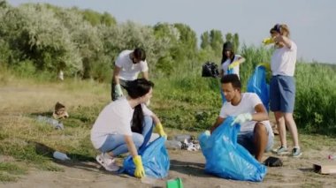 Güneşli bir günde bir grup çok ırklı gönüllü çöpleri temizliyor plajı temizliyor mavi plastik torbalardaki çöpleri topluyorlar.