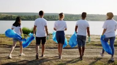 Sahilde çok ırklı büyük bir grup gönüllü olarak yürüyor koruyucu eldivenler giyiyorlar ve plajdan çöp ve plastik toplamak için büyük mavi plastikler tutuyorlar.