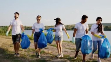 Çok ırklı gönüllüler sahildeki büyük bir gölün yanında karizmatik genç bir grup çöpleri temizledikten sonra plastik mavi torbaları alıp kameranın önünde yürümeye başlarlar.