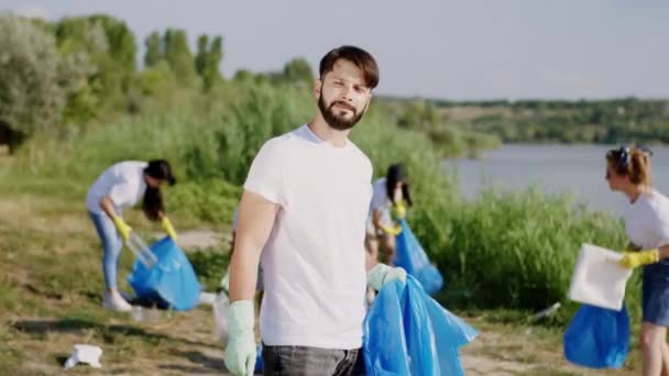 Närbild till kameran killen med en blå plastpåse städa upp skräp från stranden med sina vänner multietniska volontärer de tar hand om efter naturen — Stockvideo