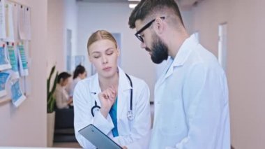 Modern hastane resepsiyonunda iki doktor birlikte hastaların geçmişlerini inceliyor. Sandalyelerde oturan bir hasta muayene için çağrılmayı bekliyor.