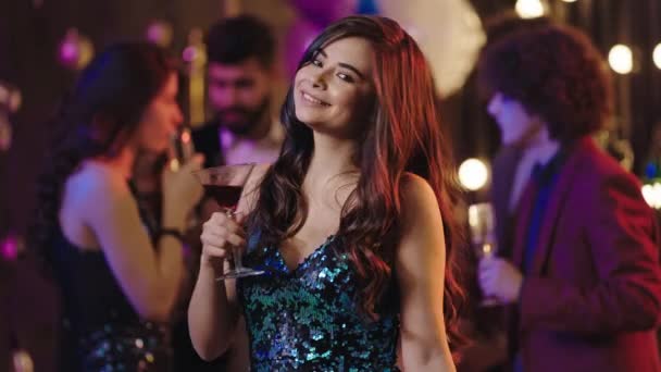 Gut aussehende junge Frau mit einem breiten Lächeln auf Geburtstagsparty mit einem Glas Getränk und Blick in die Kamera und bewegt sich langsam — Stockvideo