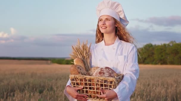 Bello con un bel sorriso bionda capelli panettiere signora in possesso di un cesto con pane fresco di fronte alla fotocamera in mezzo al campo di grano — Video Stock