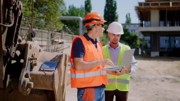 Middle-aged foreman and engineer guy with a protective uniform using the laptop to check the construction of building they have a discussing concentrated. 4k — Stock Video