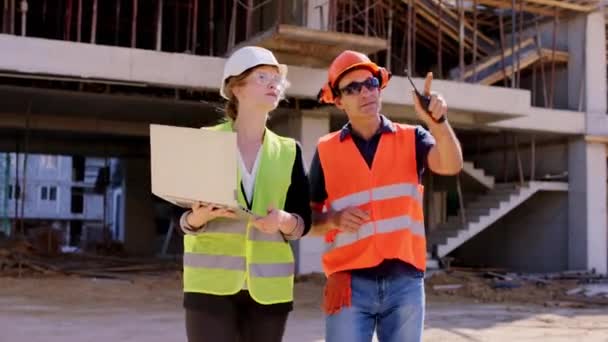노트북을 든 젊은 엔지니어 숙녀가 노인 옆을 걷고 있습니다. 그들은 안전 헬멧과 고글을 쓰고 있는 건설 현장의 계획을 분석하고 있습니다. — 비디오
