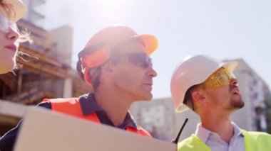 Kameranın önünde ustabaşı ve ekibi birlikte dalış uzmanları inşaat alanındaki problemleri analiz ederken güvenlik ekipmanları giyiyorlar.