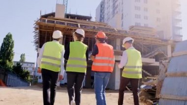 İnşaat alanındaki çeşitli etnik çeşitlilikteki uzmanlardan güvenlik kaskı takarak birlikte çalışma planını analiz eden bir grubun arkasından video alıyoruz. 4k