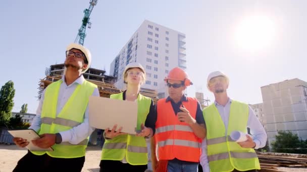 In de voorkant van de camera jonge ingenieurs multi-etnische en de voorman van middelbare leeftijd lopen door de bouwplaats en analyseren van het plan van het gebouw samen met de voorman met behulp van rantsoen om — Stockvideo