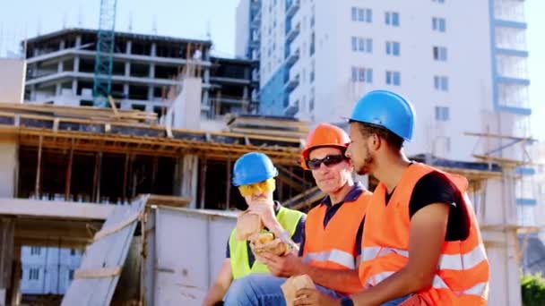 Kameraya yakın olan çok ırklı karizmatik inşaat işçileri inşaat sahasının ortasında öğle yemeği yiyorlar ve oturup sandviç yemeye başlıyorlar. 4k — Stok video