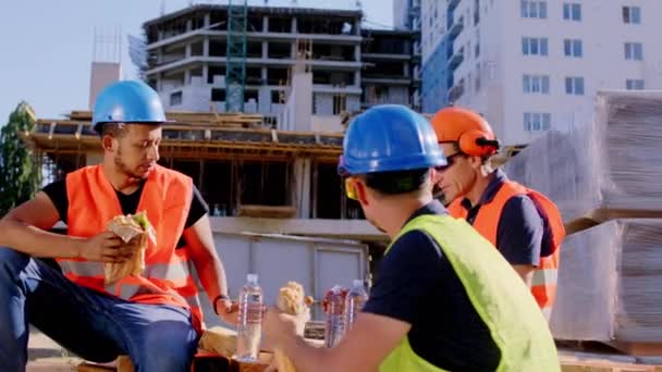 İnşaat alanında güvenlik miğferi ve gözlüklü çok ırklı işçiler biraz sandviç yiyip sohbet ediyorlar. — Stok video