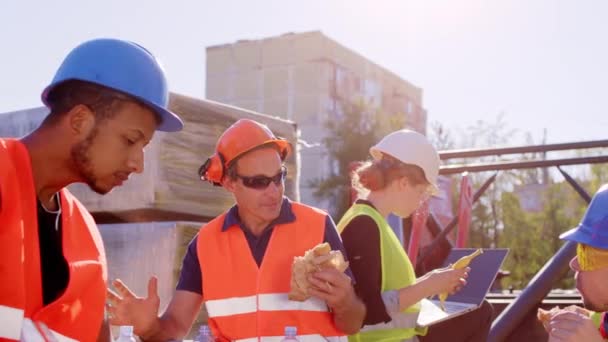 An einem sonnigen Tag auf der Baustelle machen verschiedene Spezialisten eine Pause beim gemeinsamen Essen und diskutieren gleichzeitig mit Schutzhelmen und Schutzbrillen. 4k — Stockvideo