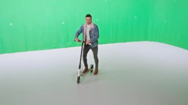 Yeşil stüdyo konsepti iyi görünümlü genç adam elektrikli scooter durağında kameranın önünde duruyor ve büyük bir gülümsemeyle poz veriyor.