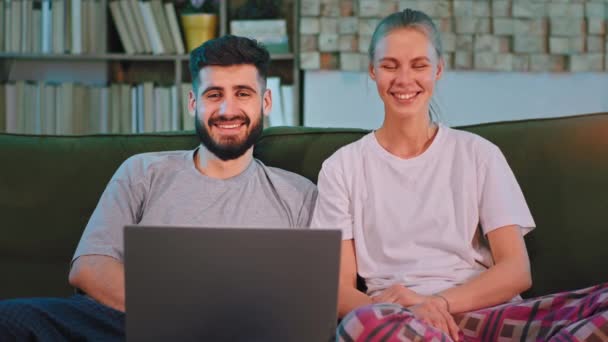 Muito carismático jovem casal em casa no sofá olhando diretamente para a câmera e sorrindo grande eles segurando um laptop ao mesmo tempo — Vídeo de Stock
