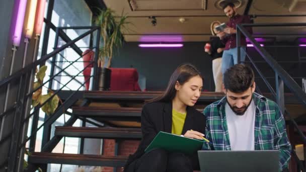 Atractiva estudiante japonesa y su colega de la universidad haciendo su proyecto juntos en el cuaderno toman notas en el mapa mientras sus colegas bajan por la escalera de la — Vídeos de Stock