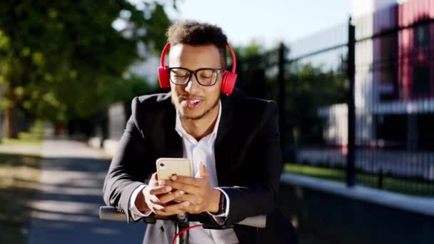 Portrait d'un homme charismatique afro-américain avec un casque faire un arrêt au milieu de la rue il prend son smartphone et analyser quelque chose portant des écouteurs rouges — Video