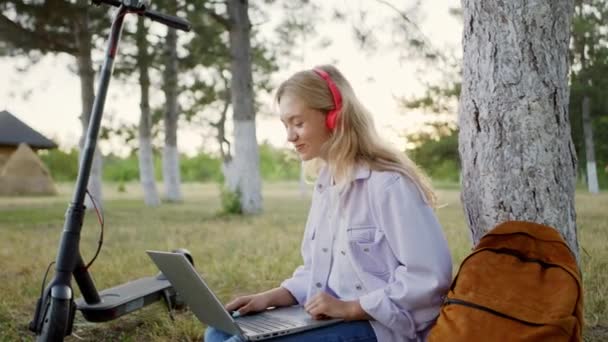 Młoda studentka z charyzmatyczną twarzą ma rozmowę kwalifikacyjną z laptopem online, a ona siedzi na trawie w środku parku w czerwonych słuchawkach. — Wideo stockowe