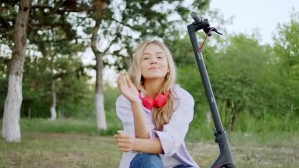 Jovem bonita com um sorriso bonito olhando direto para a câmera e posando enquanto faz uma parada com sua scooter elétrica — Vídeo de Stock