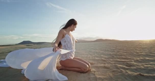 Сексуальная и красивая дама посреди пляжа сидит и держит свое длинное белое платье, которое она наслаждается временем на закате — стоковое видео