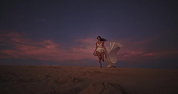 Hermosa mujer joven caminando a través de la arena romántica playa que sostiene su vestido blanco largo romántico. 4k — Vídeos de Stock