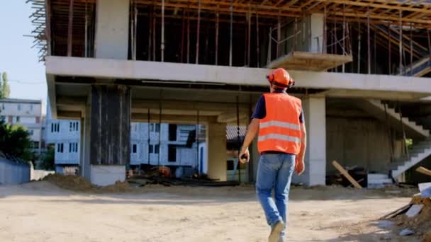 En el sitio de construcción grande caminar capataz en un equipo de seguridad para el sitio de construcción tomar video desde la parte posterior — Vídeos de Stock