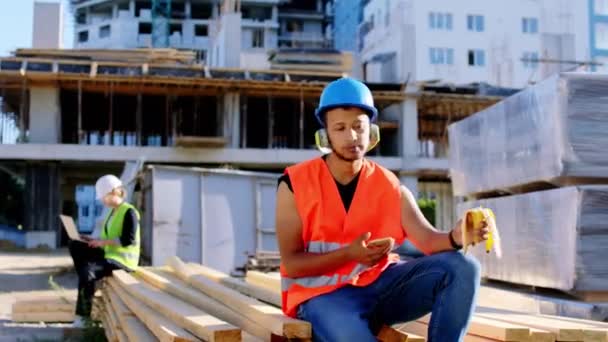 Bien parecido joven afroamericano tener un descanso en el sitio de construcción que se come un plátano y escuchar música de los auriculares. 4k — Vídeos de Stock