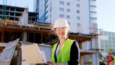 Katmanlı bir gülümseme ile inşaat alanında güzel bir mühendis kadın kameranın önünde poz veriyor ve dizüstü bilgisayarını tutuyor ve gözlük takıyor ve güvenlik kaskı takıyor.