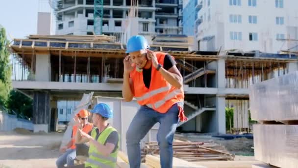 Büyük bir inşaat sahasında inşaat işçileri mola zamanında dans ediyor ve mutlu hissediyorlar. Hepsi kask takıyor ve koruyucu üniforma giyiyorlar. — Stok video