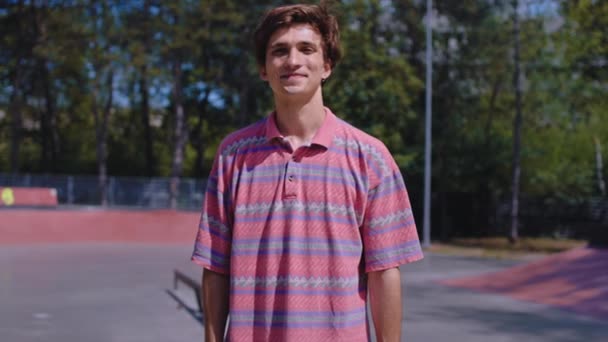 Sonriendo lindo chico carismático joven delante de la cámara en un moderno parque de skate tiene un gran estado de ánimo mirando directamente a la cámara — Vídeos de Stock