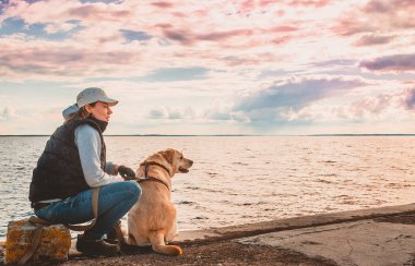 Bir kız rıhtımda Labrador Retriever 'la oturuyor ve manzaraya hayran. Köpekle akşam gezintisi