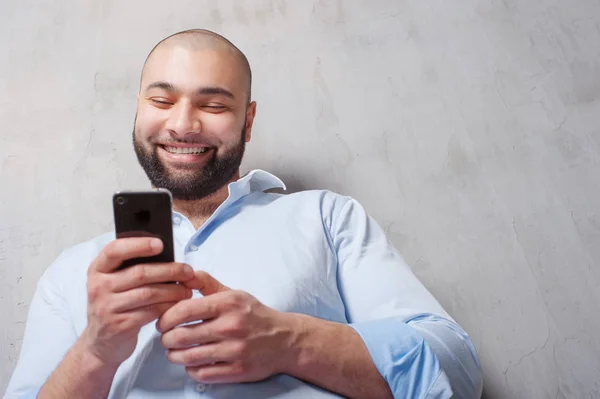 Handsome young man in shirt using mobile phone and smiling while standing against grey wall.