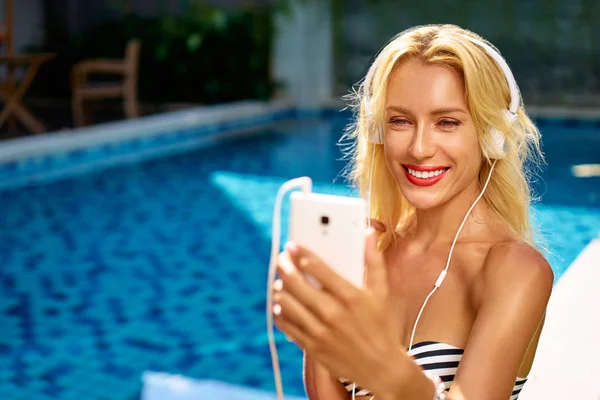漂亮的年轻女子在比基尼使用智能手机在游泳池附近的耳机 — 图库照片