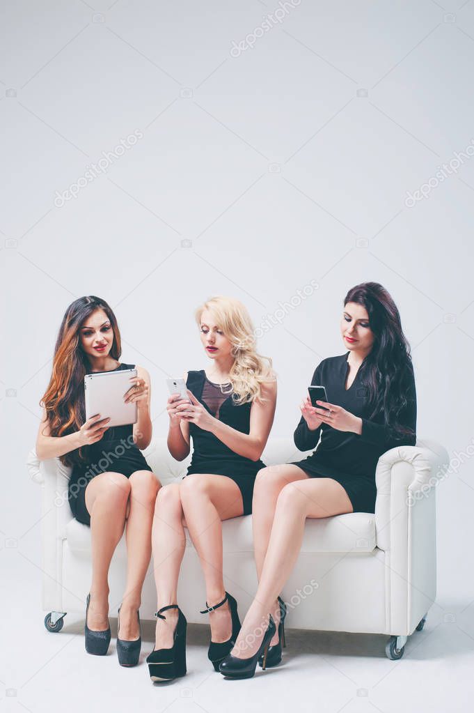 Three gorgeous young women using digital gadgets while  sitting on white sofa.