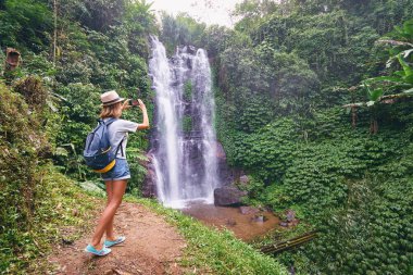 Sırt çantası alarak fotoğraf onun Smartphone cep telefonu ile büyük şelale ile şapkalı genç kadın.