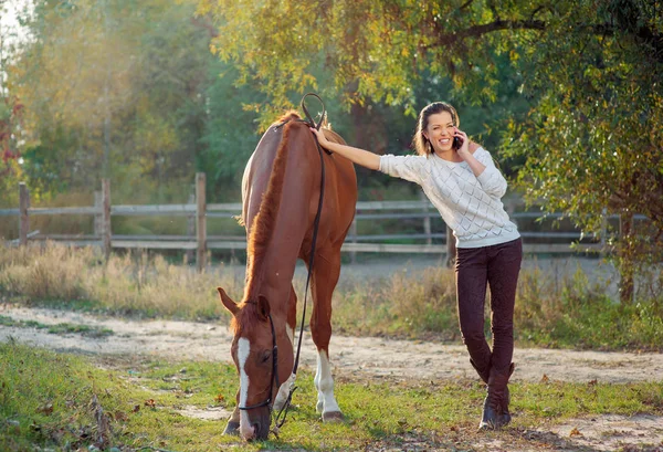 Attractive young woman talking by phone while walking with horse outdoors.