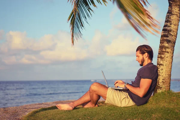 在棕榈树下热带海滩上的笔记本电脑工作的年轻人 — 图库照片